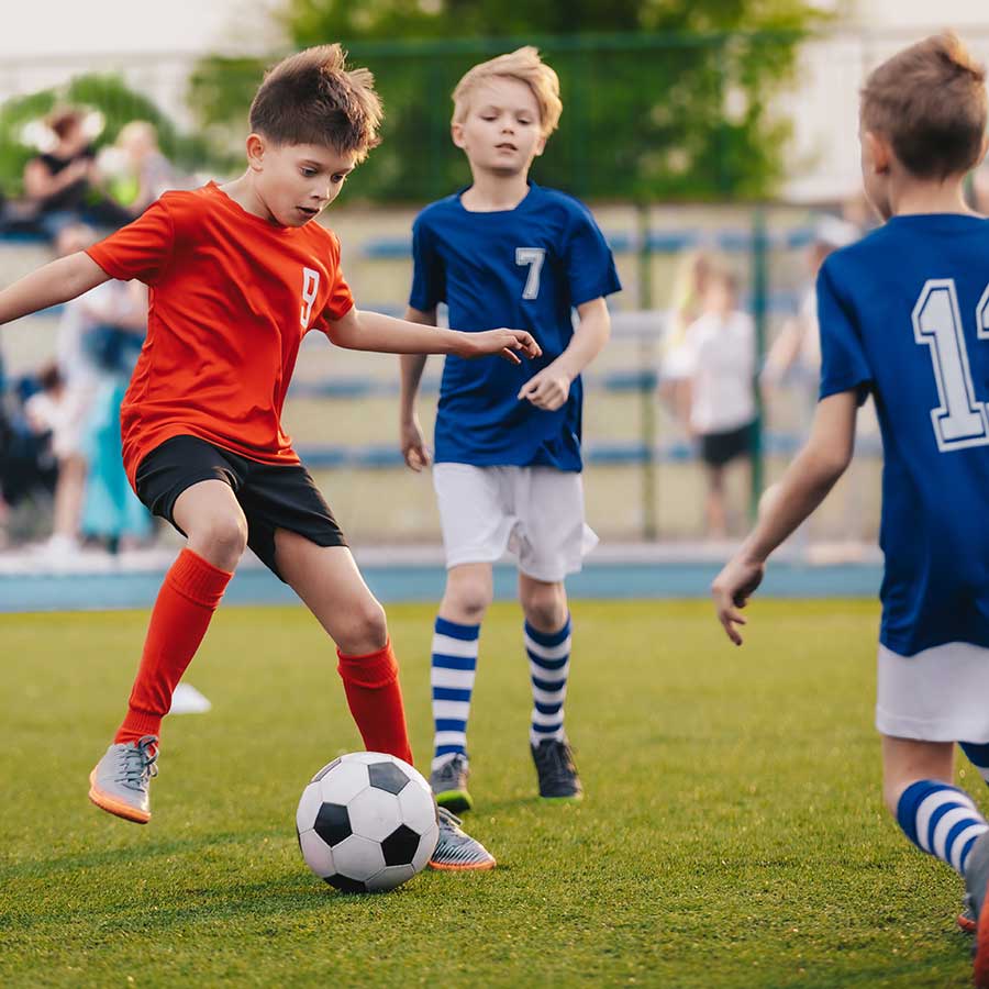 Three boys play soccer on a field; one in a red jersey controls the ball while two in blue jerseys approach to defend.