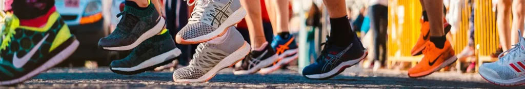 Close-up of several runners' legs and colorful running shoes in motion on a street, with a blurred background and orange barriers.