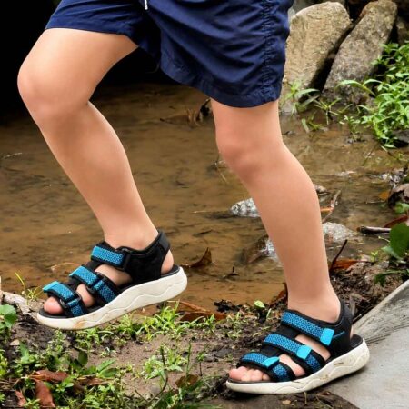 A child in blue shorts and 'Arch-Up' sandals - Black/Blue steps near a muddy puddle and rocks outdoors.
