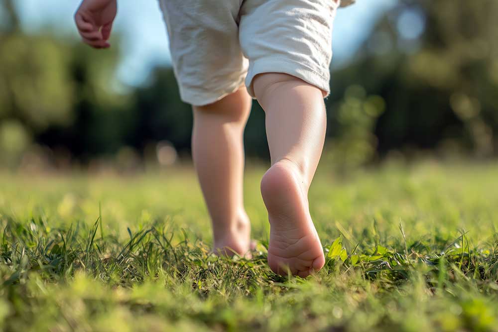 A toddler walks barefoot on green grass outdoors, with sunlight in the background.