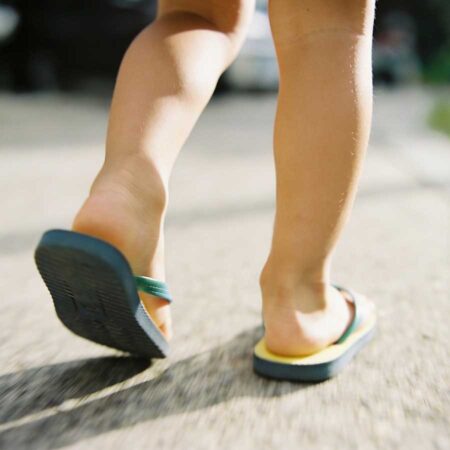 Close-up of a child's legs and feet walking outdoors on a sidewalk, wearing flip-flops.